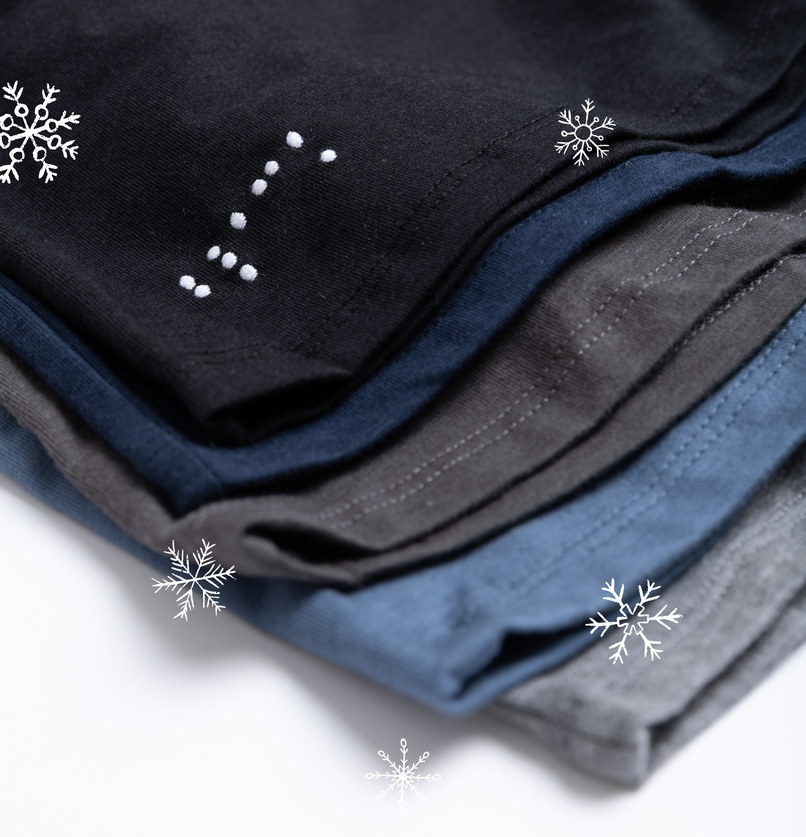 Stack of folded black, blue, and gray shirts with snowflake overlays on a white background. The top shirt features the work black in braille 