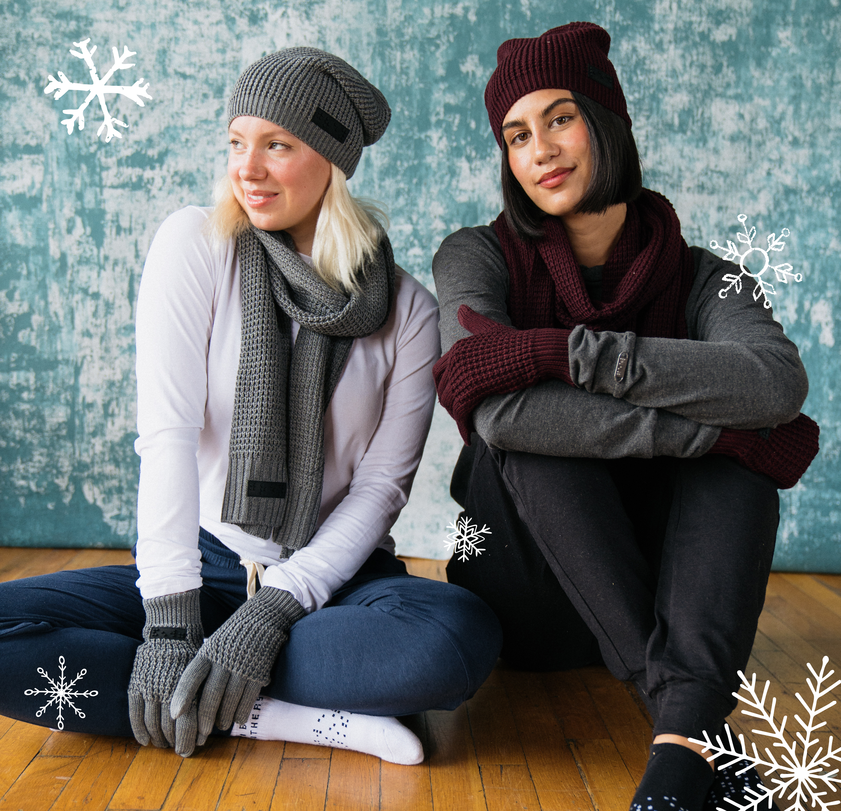 Two women wearing two blind brothers chunk knit beanies, scarves, and gloves, along with two blind brothers apparel. With a textured blue wall and snowflake overlays in the background.